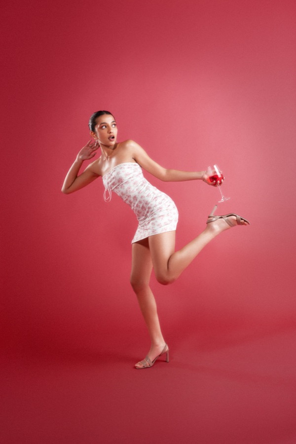 Commercial campaign — playful kicked-up pose with a wine glass on crimson backdrop